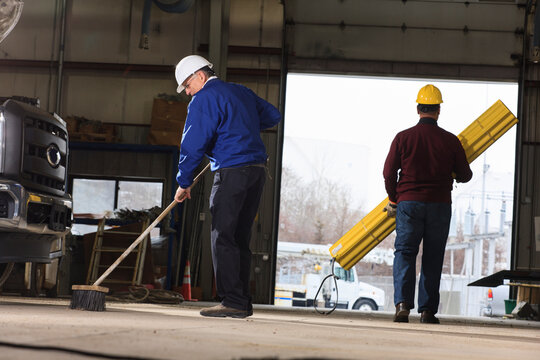Man sweeping workshop while other carrying supplies