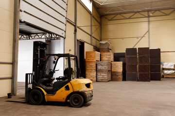Yellow forklift truck parked inside industrial warehouse next to open gate. Forklift loader vehicle near stacked wooden boxes, logistics distribution storage hub property business background © Krystsina