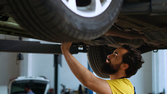 Certified repairman underneath vehicle on overhead lift in garage, checking parts. Specialist using work light to make sure automotive underbody is in proper condition, doing close examination.