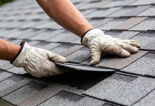 A close-up view of a worker's gloved hands repairing gray asphalt shingles on a residential roof.