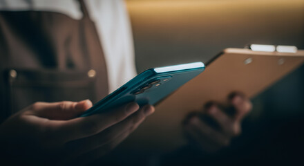Fototapeta premium Worker in an apron holding a smartphone and clipboard representing digital business management and workflow efficiency.