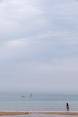 Fototapeta premium Calm beach with lone woman and distant paddle boarders.Calm beach scene with a lone woman at the shoreline and distant paddle boarders on a misty sea under a soft cloudy sky.
