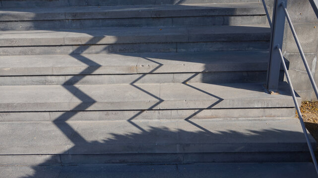 Sharp geometric zigzag shadows cast by a metal railing fall across outdoor concrete stairs under bright sunlight.