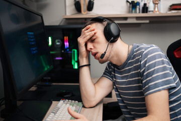 A young male gamer wearing a headset expresses frustration and fatigue, holding his head in disappointment while playing an intense online multiplayer game at his computer desk. © Dusko