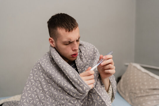 A sick young man wrapped in a blanket checks his temperature with a digital thermometer while suffering from flu or fever in bed at home.