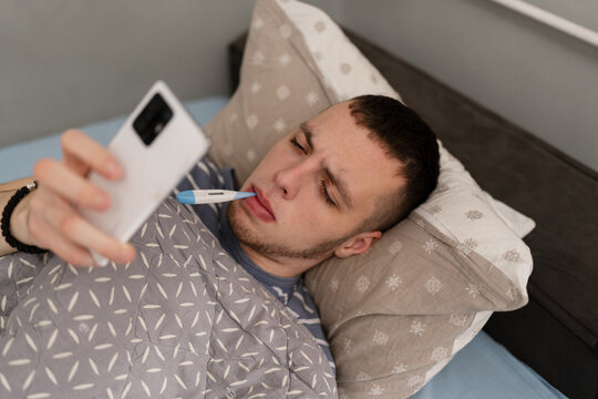 A sick young man lies in bed with a thermometer in his mouth, looking at his smartphone to check symptoms or contact a doctor during a cold or flu infection.