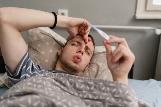 A sick young man lies in bed suffering from a high fever, checking his temperature with a digital thermometer at home during flu season.