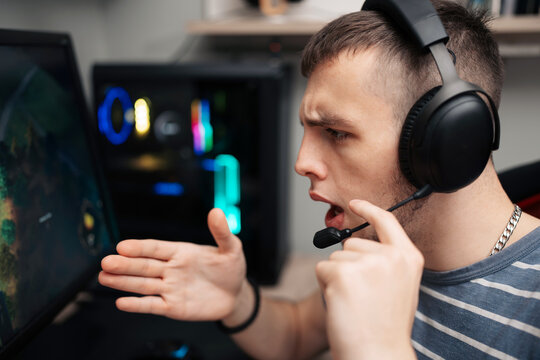 A young male gamer wearing a headset reacts intensely while playing a competitive computer video game with a custom PC rig in the background.
