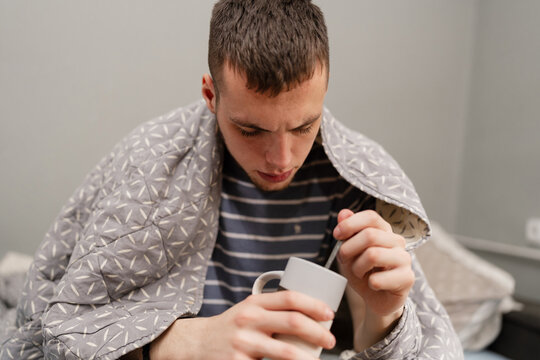 A sick young man wrapped in a warm blanket sits at home holding a mug of tea while suffering from flu symptoms.