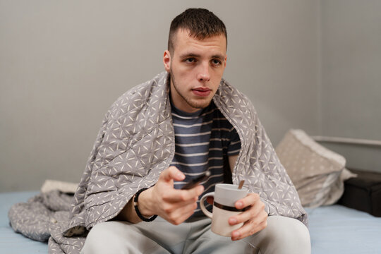 A young man feeling unwell with the flu sits in bed wrapped in a blanket, holding a mug of warm tea and a remote control while recovering at home.