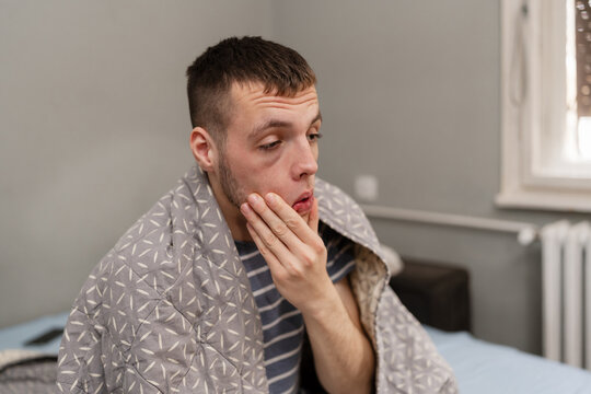 Sick young man wrapped in a blanket sitting on his bed, suffering from seasonal flu, fever, or a cold at home.