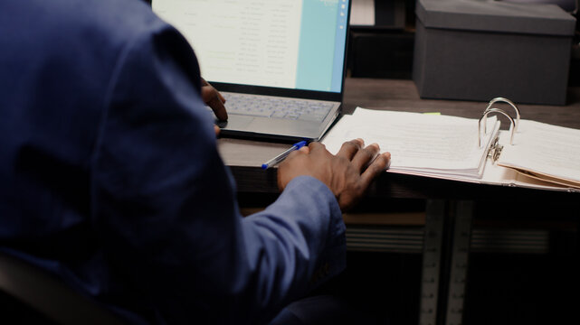 Detective captured in midst of investigation, analyzing records and evidence, with detailed view of policeman at desk with laptop and case files opened for cross-referencing suspect profile.