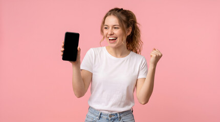 Fototapeta premium young cute woman in white t-shirt shows blank smartphone screen and rejoices in victory on pink isolated background, the winner girl advertises copy space on the phone, mockup