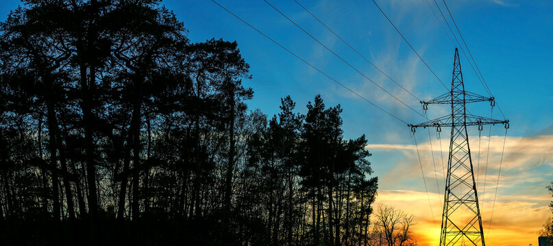 Silhouette of high-voltage power line and forest trees against blue evening sunset sky. Industrial landscape.