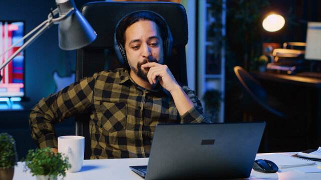 Cheerful man relaxing at home by watching TV show on laptop using headphones, happy person enjoying leisure time in neon lit apartment at night, consuming entertainment media content.