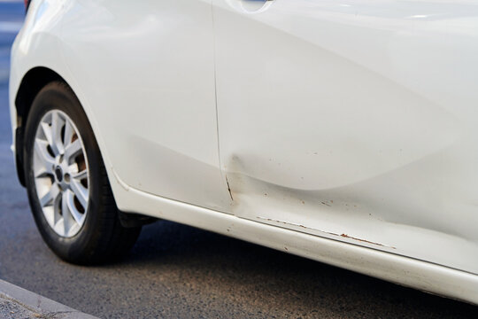 Scraped and dented white automobile door requiring fix at garage body shop, minor rust formation on metal edge from roadway scratch hazard. Lower door dent and surface paint scratches. Selective focus