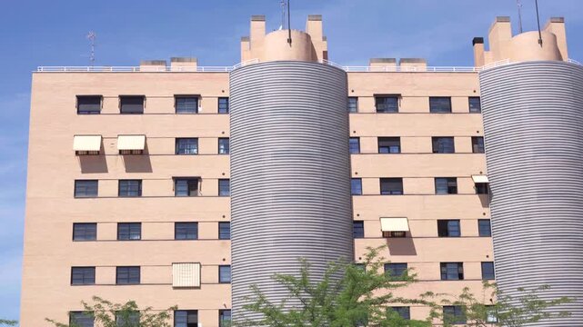 Modern residential building with two tall cylindrical towers. Light beige facade with many rectangular windows. Some windows have white awnings or balconies. Green trees at bottom frame the scene