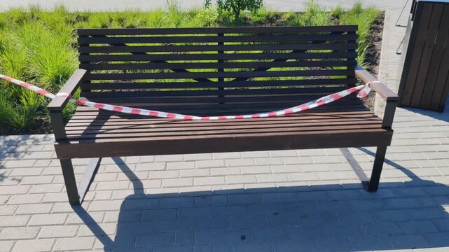 Wooden park bench blocked off with red and white caution tape after fresh painting, restricting access in public space, urban maintenance, sunny day, green bushes background