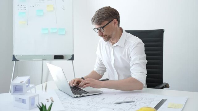 Male architect in modern office creating architectural design blueprint on paper and working on laptop, developing new building project with models on white desk