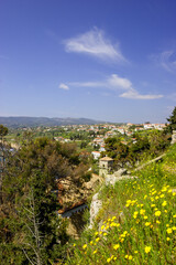 Obraz premium A scenic view of a coastal town with red-tiled roofs and white buildings, nestled among green hills under a bright blue sky with wispy clouds. 