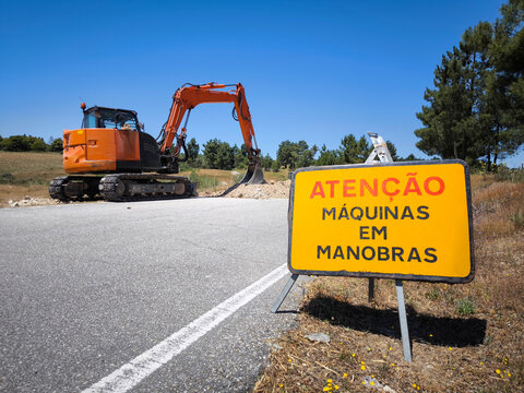 Placa de sinaliza&ccedil;&atilde;o com a descri&ccedil;&atilde;o em portugu&ecirc;s "Aten&ccedil;&atilde;o: M&aacute;quinas em Manobras" com uma retro-escavadora a realizar trabalhos de escava&ccedil;&atilde;o e abertura de valas para saneamento na berma de uma estrada