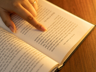 Fototapeta premium Close-up of a woman's hands holding an open book with clearly visible text pages, shot in warm cozy indoor lighting with a soft glowing table lamp in the background, evoking reading and knowledge. 