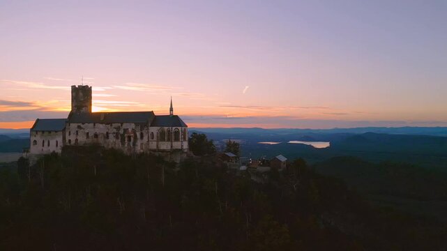The camera captures Bezdez Castle at sunset. The sun dips below the horizon, casting warm light over the castle and surrounding hills.