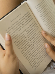 Fototapeta premium Close-up of a woman's hands holding an open book with clearly visible text pages, shot in warm cozy indoor lighting with a soft glowing table lamp in the background, evoking reading and knowledge. 