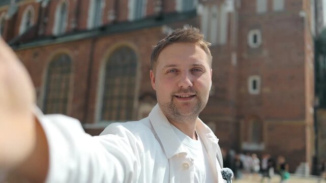 Medium shot of a happy young caucasian male tourist vlogging and recording a selfie video with classic old town architecture in the background.