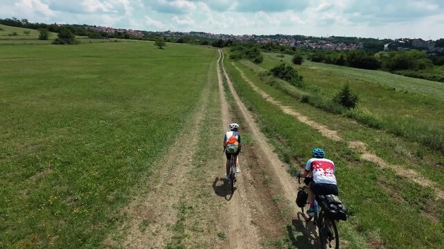 Drone view of two cyclists riding gravel bicycles along a dirt track through open countryside fields during a bikepacking adventure