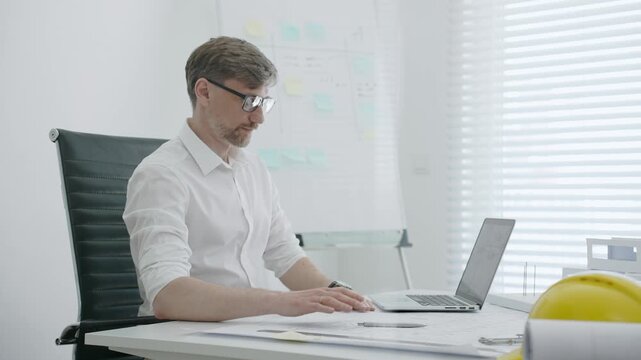 Male architect in modern office creating architectural design blueprint on paper and working on laptop, developing new building project with models on white desk