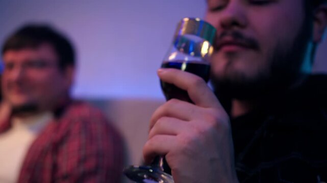 Bearded caucasian man holds, examines glass of rich red wine and drinks alcohol. Company of friends in festive atmosphere. Adult man savoring luxurious glass with beverage in evening scene in home