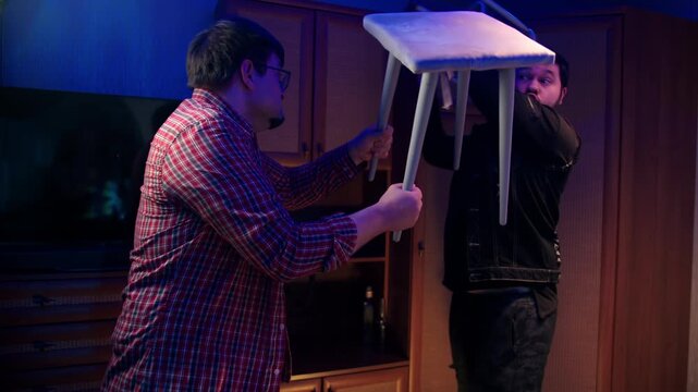Intense scene where drunk agressive caucasian man lifts stool and strikes during dispute. Men are in conflict raise seat during heated confrontation in room. Antisocial behavior after drinking alcohol
