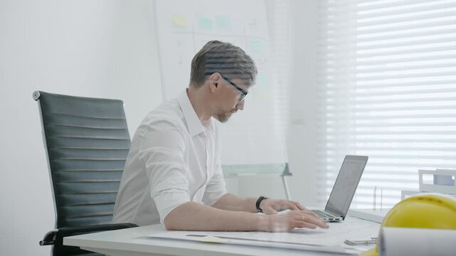 Male architect in modern office creating architectural design blueprint on paper and working on laptop, developing new building project with models on white desk