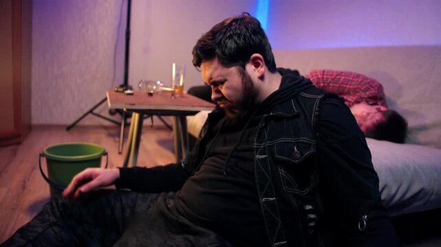 Man in drunken state vomits into green bucket close by caucasian friend on couch in apartment. Scene shows intoxicated bearded black-haired man that sits and feels nauseous after drinking alcohol