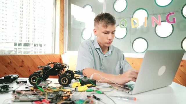 Smart student working on laptop while analysis car model construction. Happy high school boy using computer to study or learn about robotic model structure on table with electronic tool. Edification