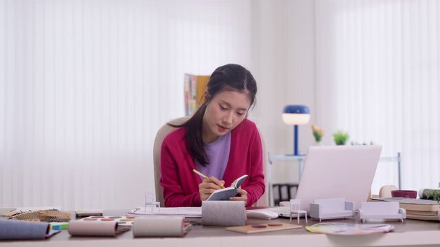 young asian woman furniture designer planning interior design project writing notes with laptop computer home office workspace creative professional studying