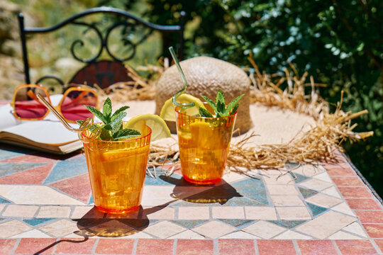 Summer refreshing lemonade drink or non alcoholic cocktail with lemon slices in orange drinking glasses on the table in the garden. Fresh healthy cold lemon beverage. Water with lemon. Summer mood.