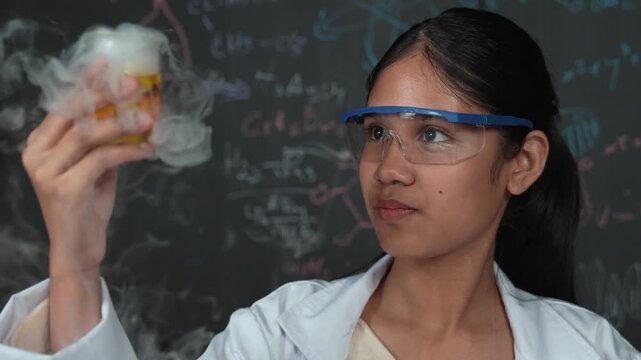 Closeup of smart high school student inspect colored solution at blackboard. Beautiful scientist holding beaker and looking at chemical liquid while standing in front of blackboard at lab. Edification