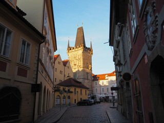 Obraz premium Sunrise illuminates the cozy streets of Mala Strana in Prague, Czech Republic. The warm golden light illuminates the historic Gothic tower and buildings, creating a magical morning atmosphere.
