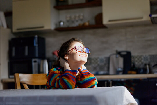 A thoughtful girl in a colorful striped sweater sits at a table in a warm home kitchen, gazing upward with a hopeful smile, conveying curiosity and calm.