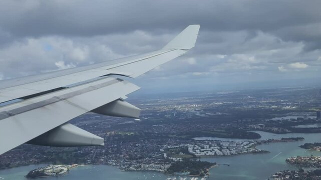 Sydney, Australia - Airplane wing view over coastal city and blue water during flight, showcasing aerial travel perspective with shoreline, clouds and urban landscape from in‑flight window.
