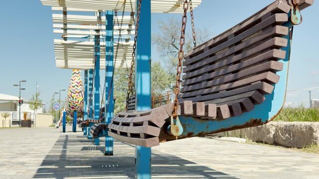 A row of slatted wooden swing benches hangs from blue posts under white shade canopies. The bright daytime setting gives a playful, nostalgic, and deeply relaxing feeling of summer leisure.