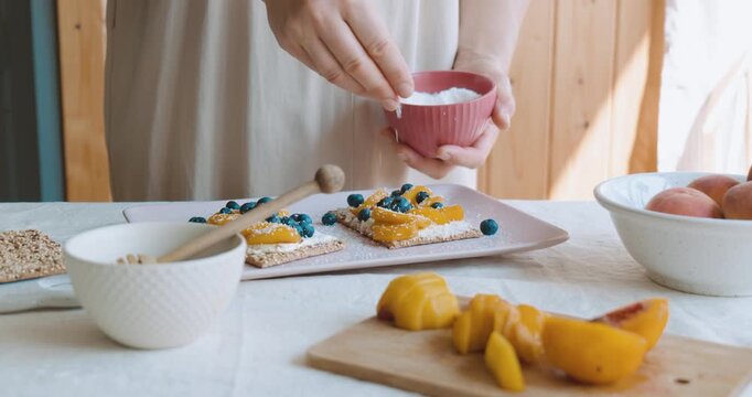Woman sprinkle with coconut flakes sweet sandwiches made with crispbread, cream cheese, blueberry, slices of peach and honey for summer picnic or brunch