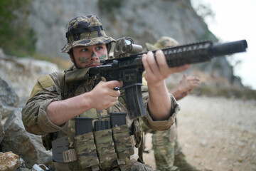 Fototapeta premium Soldier in camouflage aiming an assault rifle in a rocky outdoor environment