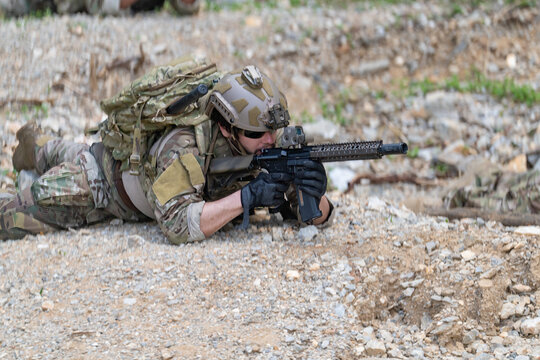 Soldier in camouflage gear aims rifle while lying prone on rocky terrain