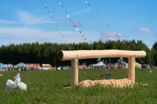 Horizontal log on a green field for the traditional bag fight competition at the Sabantuy folk festival, with colorful flags and a blue sky in the background.
