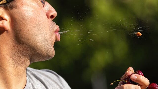 Man in profile spitting a cherry pit while holding ripe cherries outdoors