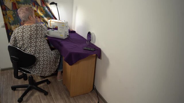 Elderly senior woman sews on sewing machine. Grandmother home craft table