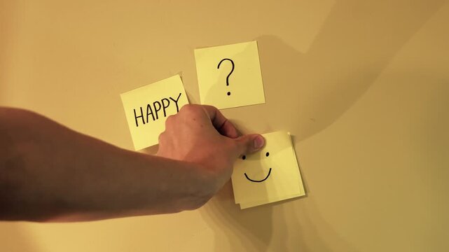 Human hand replacing a sticky note with a sad emoticon for one with a happy smiley face, representing a choice for happiness, positivity, and improved mental health and well being